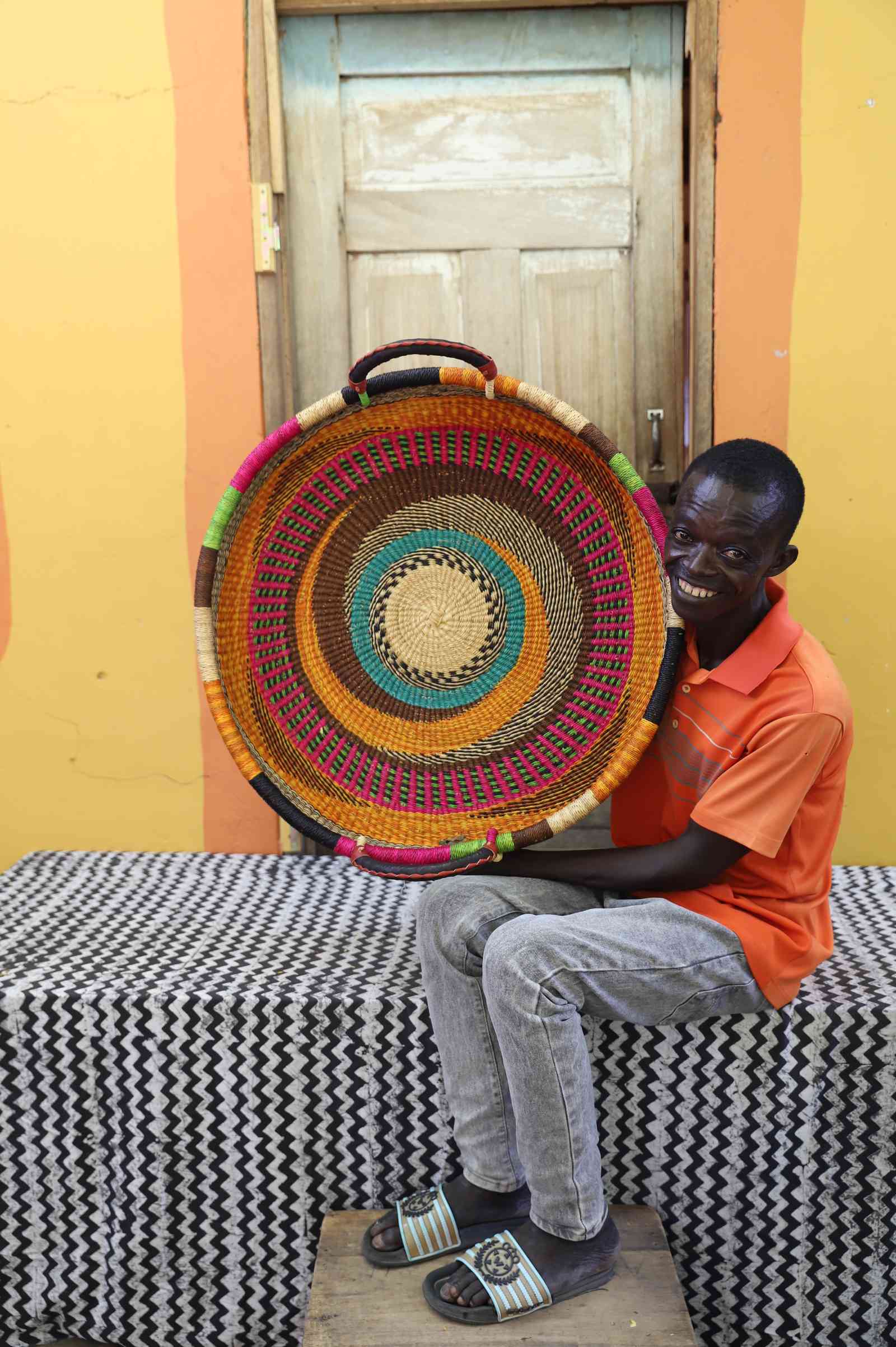 Large handwoven Bolga wall tray disc with bright multicolour bands, held by a seated man in orange.