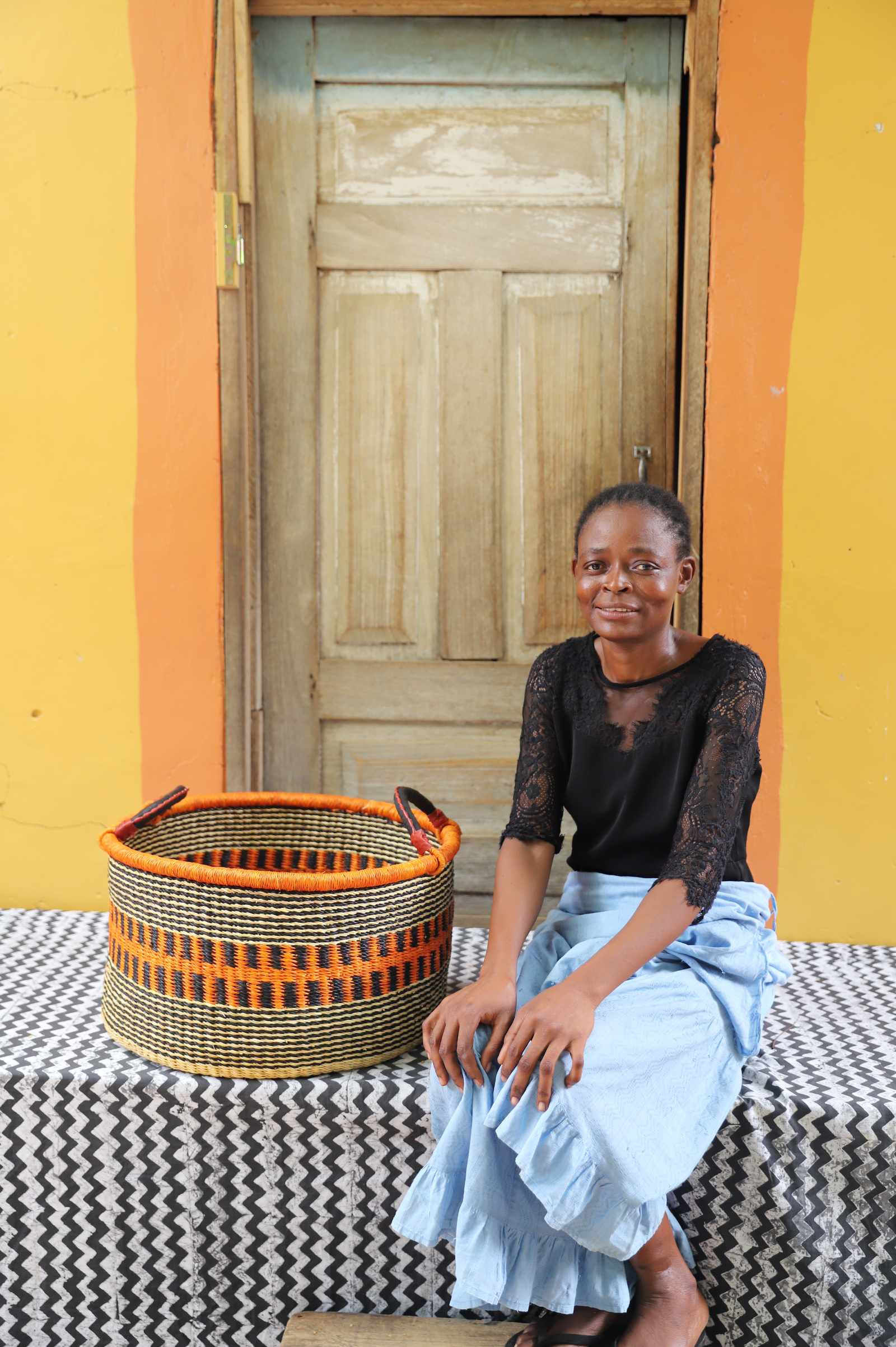 Handwoven Bolga storage basket in natural straw with an orange striped band, styled beside a seated woman.