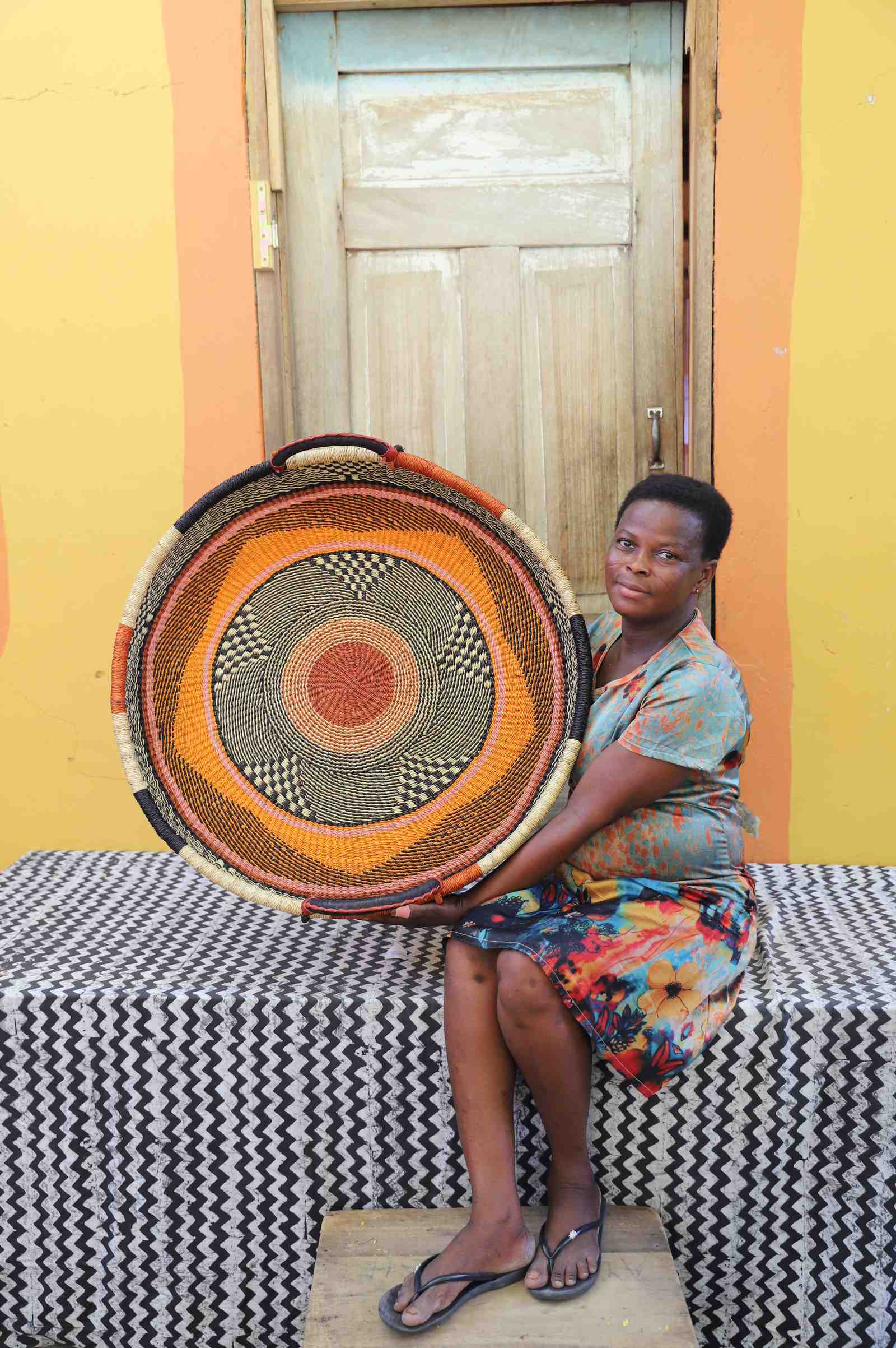 Large handwoven Bolga wall tray disc with orange, black and red bands, held by a seated woman in floral.