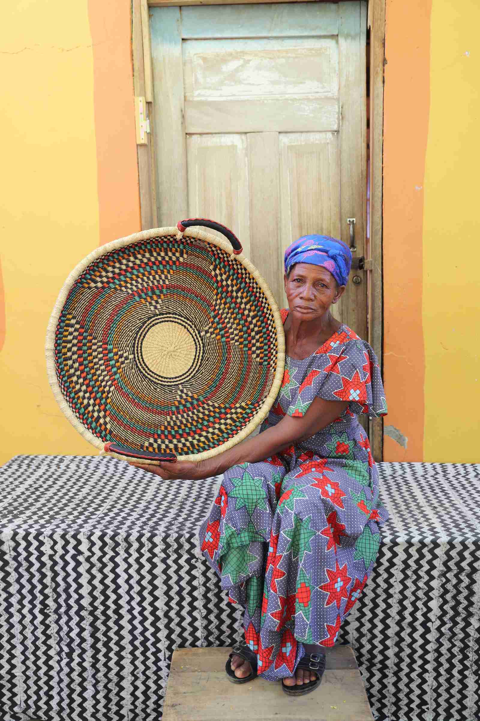 Medium handwoven Bolga wall tray disc with fine dark concentric bands, held by a seated woman in a printed dress.