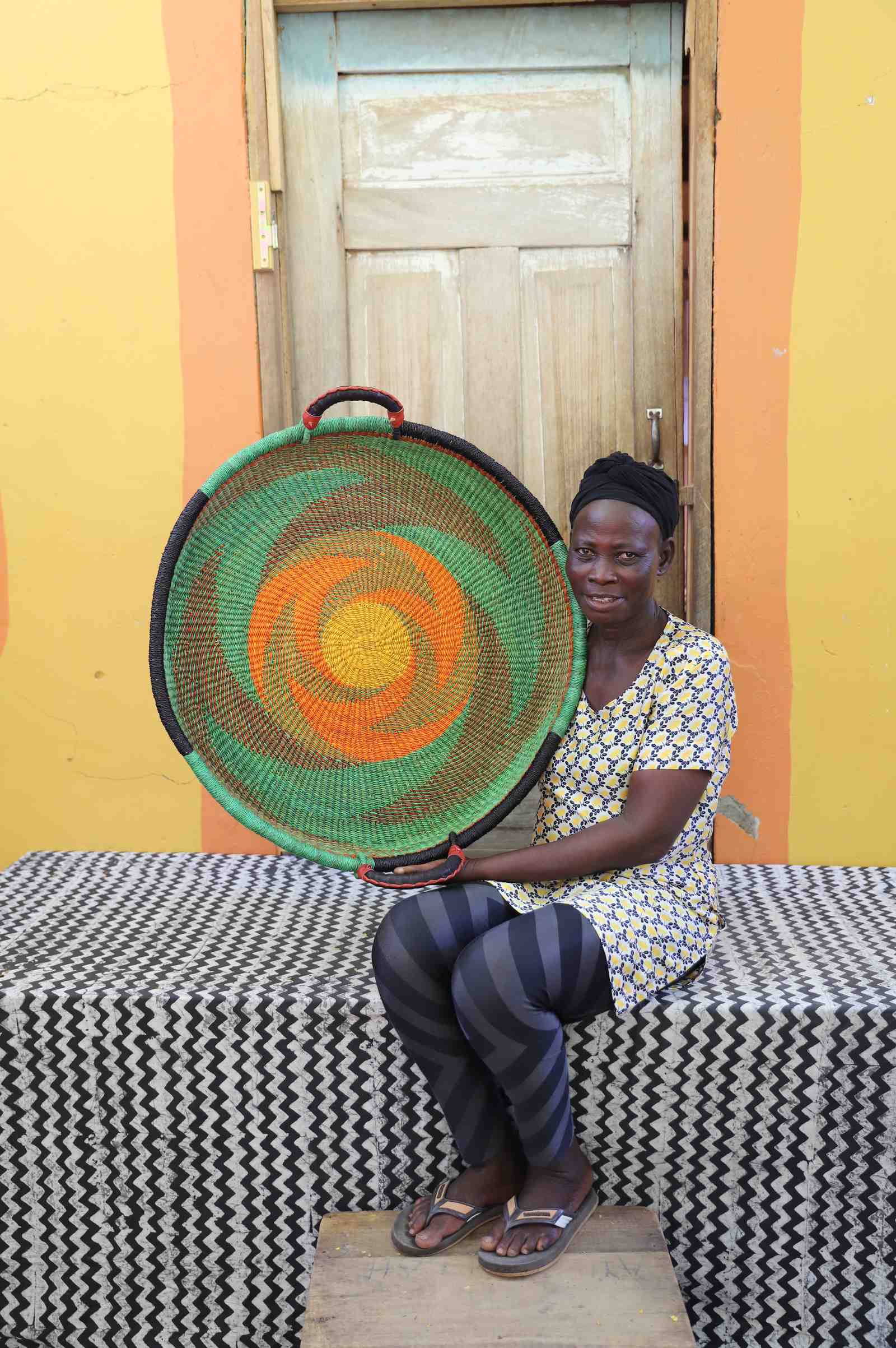 Medium handwoven Bolga wall tray disc with green body and orange swirl, held by a seated woman in white.