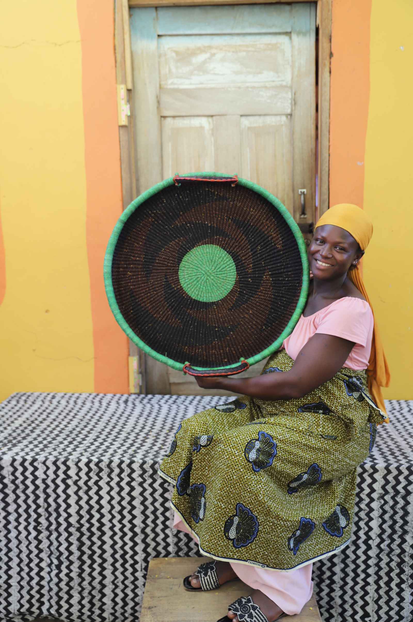 Medium handwoven Bolga wall tray disc in dark straw with green rim and centre, held by a seated woman in pink.