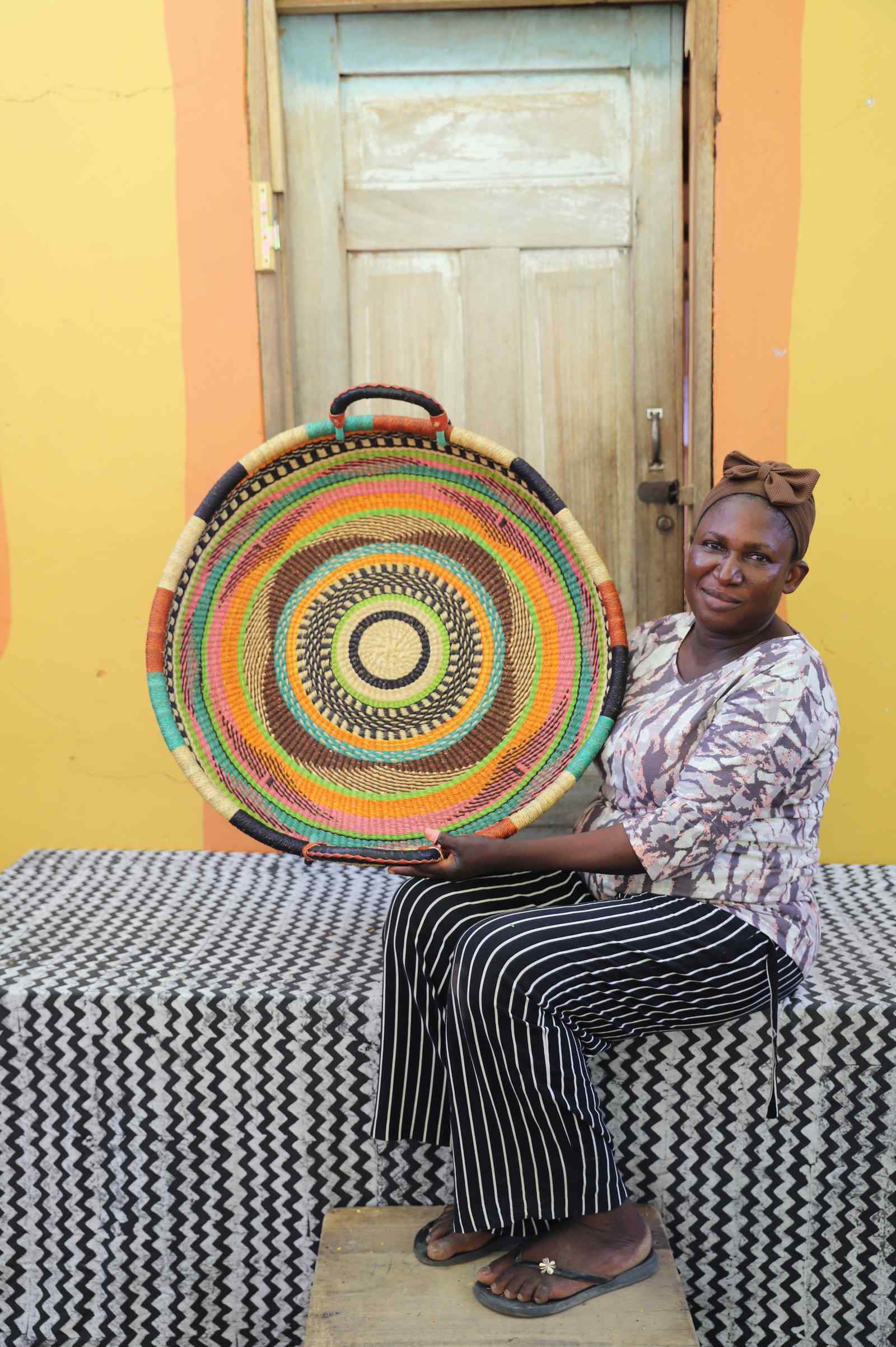 Large handwoven Bolga wall tray disc with multicolour concentric rings, held by a seated woman.