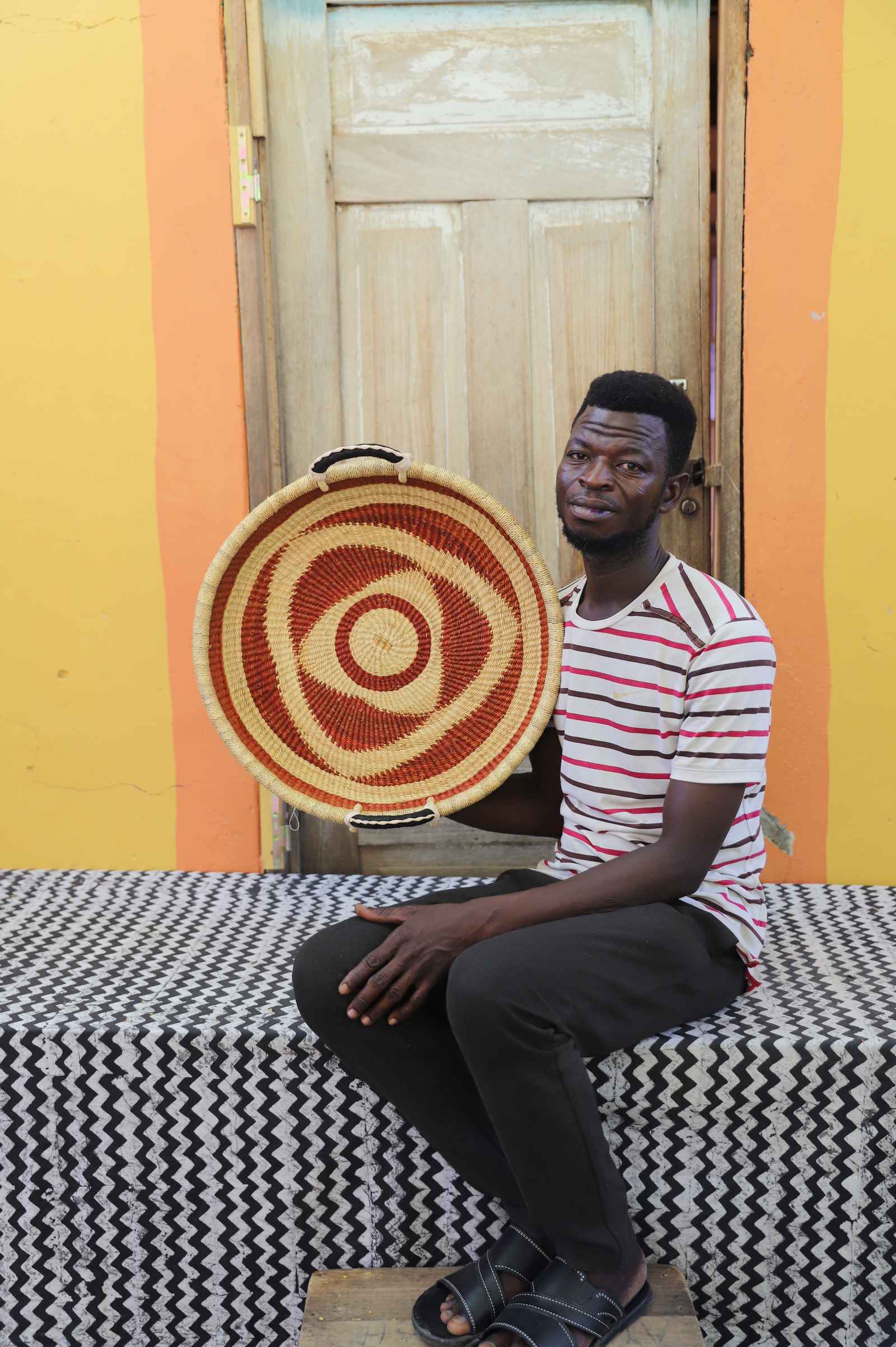 Handwoven Bolga wall tray disc with rust-red bullseye rings, held by a seated man in a striped top.