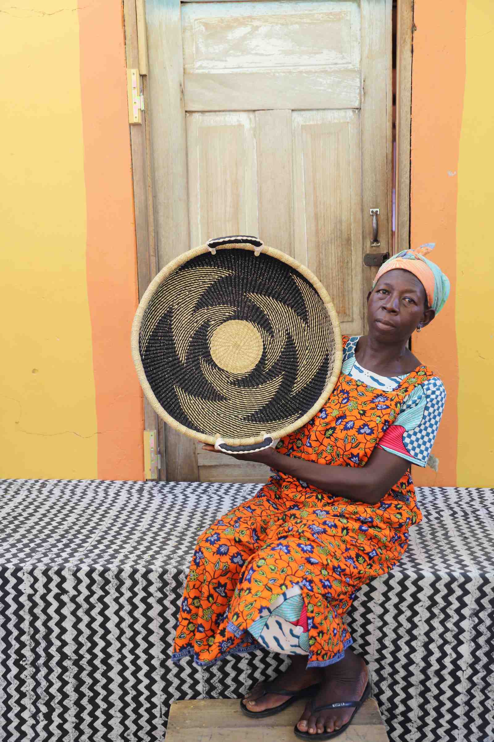 Handwoven Bolga wall tray disc with dark pinwheel swirl, held by a seated woman in an orange dress.