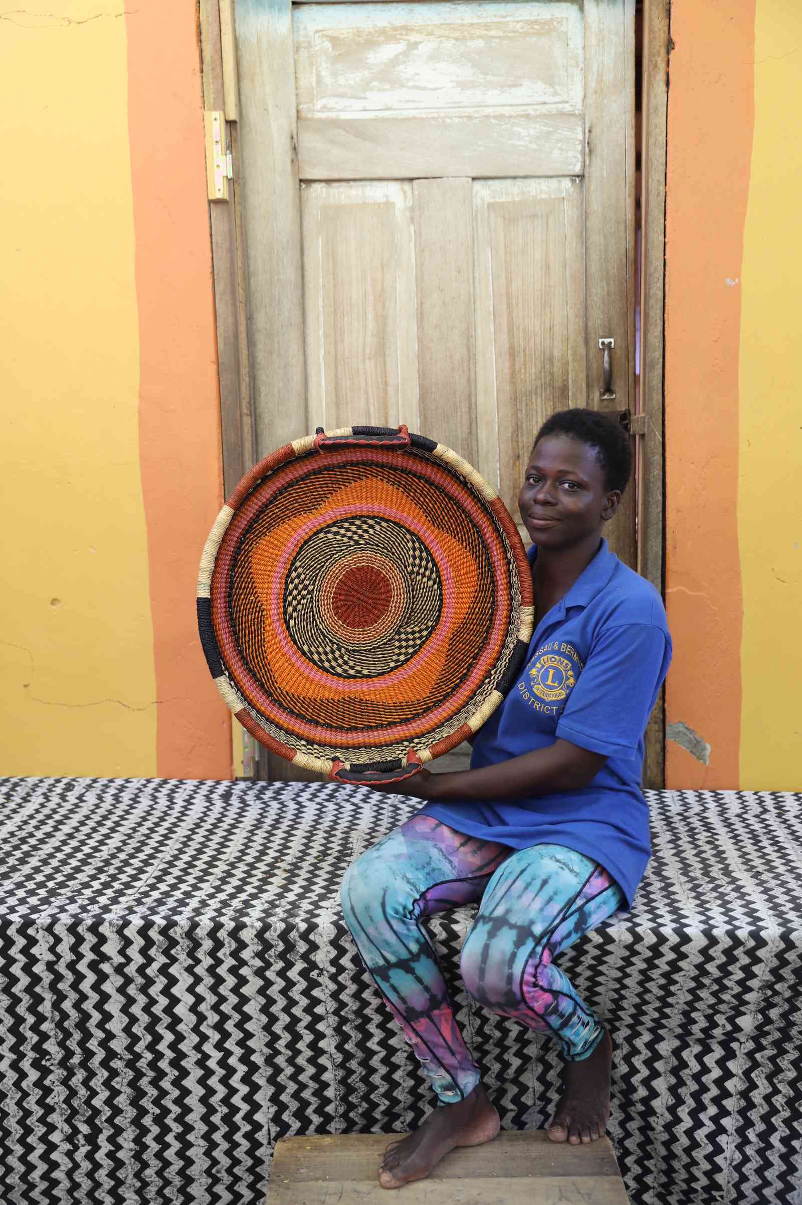 Handwoven Bolga wall tray disc in rust-orange with black rings, held by a seated woman in blue.