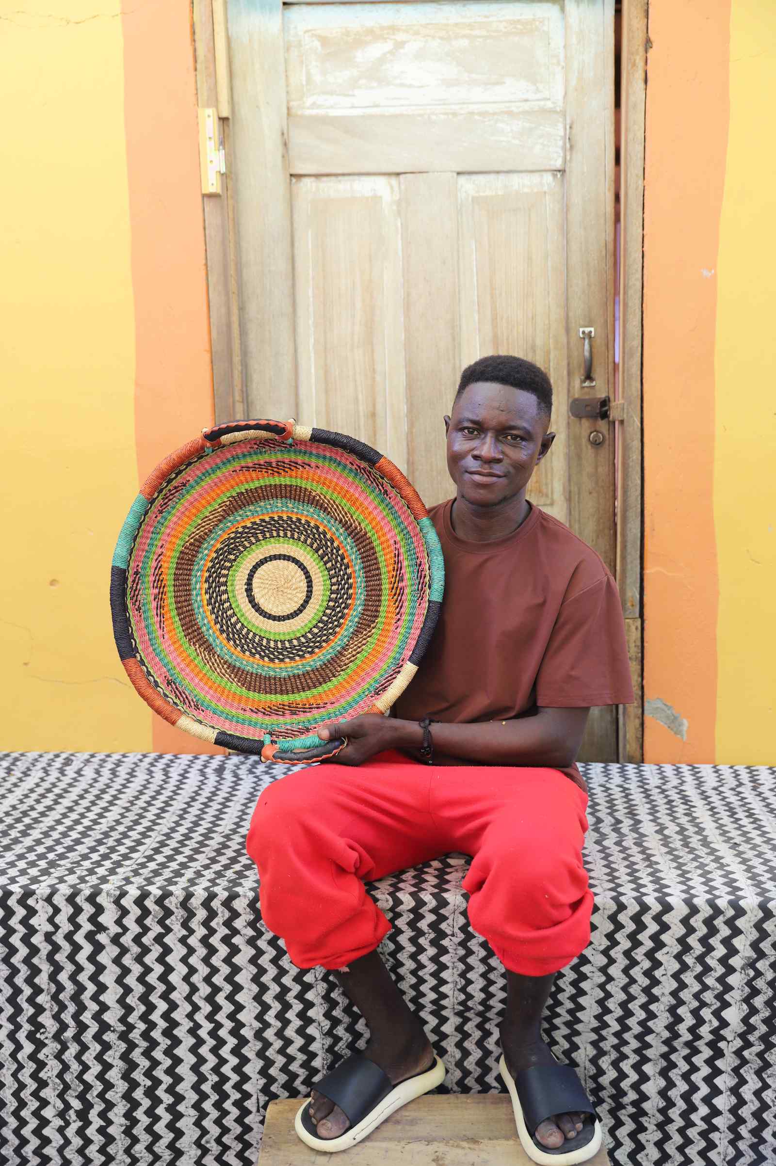 Handwoven Bolga wall tray disc with concentric multicolour rings, held by a seated man in red.