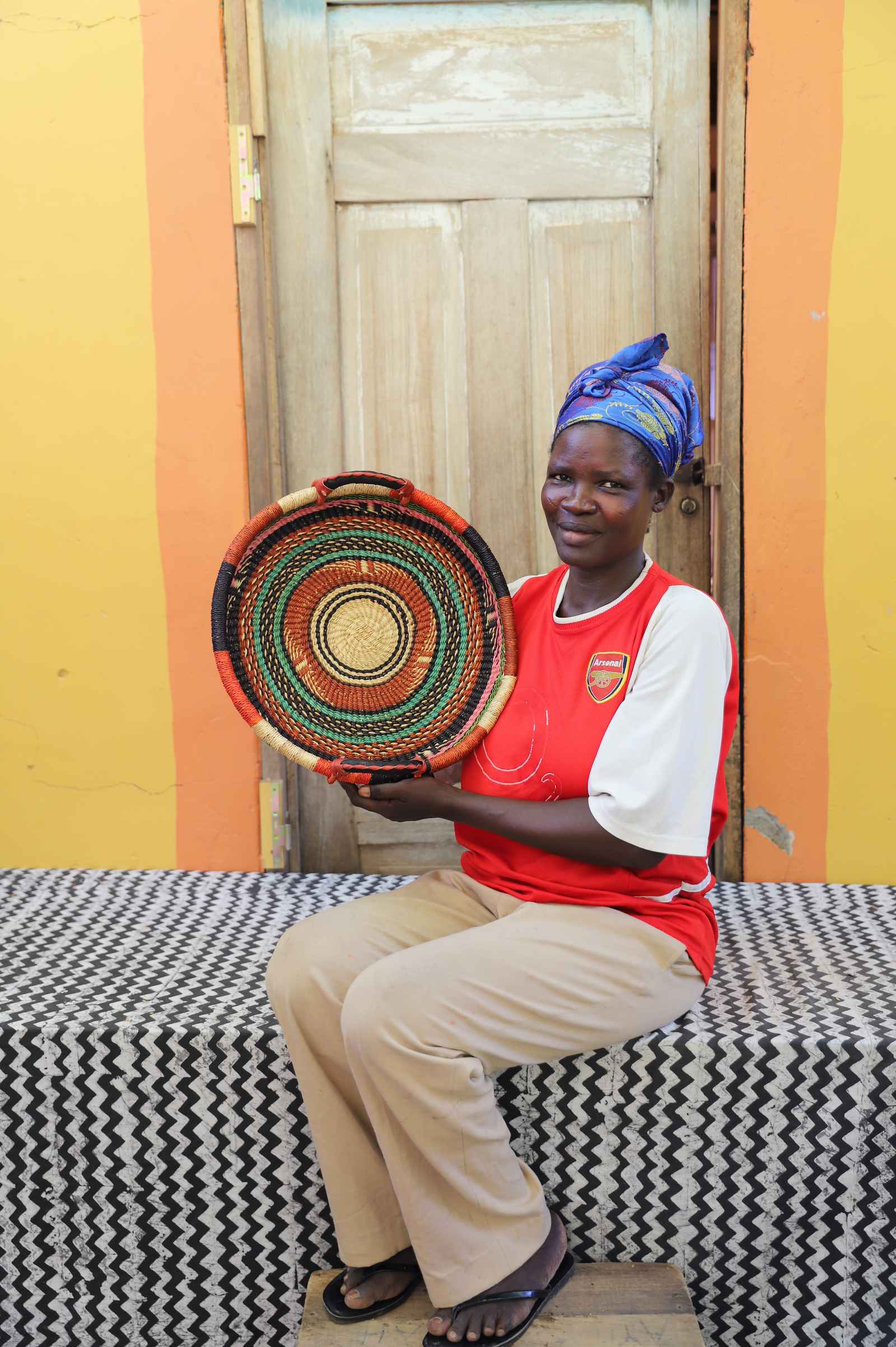 Handwoven Bolga wall tray disc in orange, black, red and green bands, held by a seated woman.