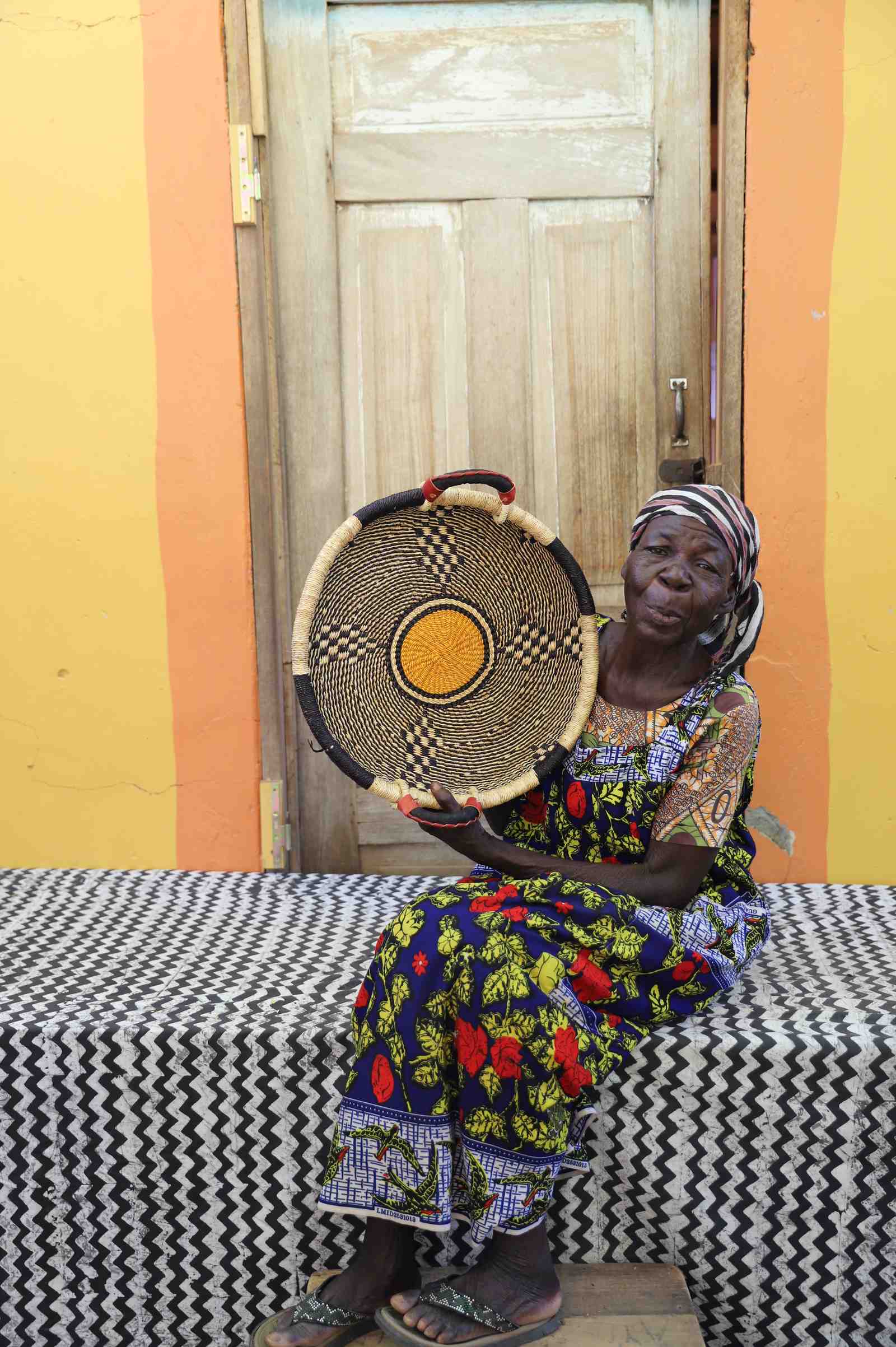 Handwoven Bolga wall tray disc in natural straw with a single yellow centre dot, held by a seated woman.