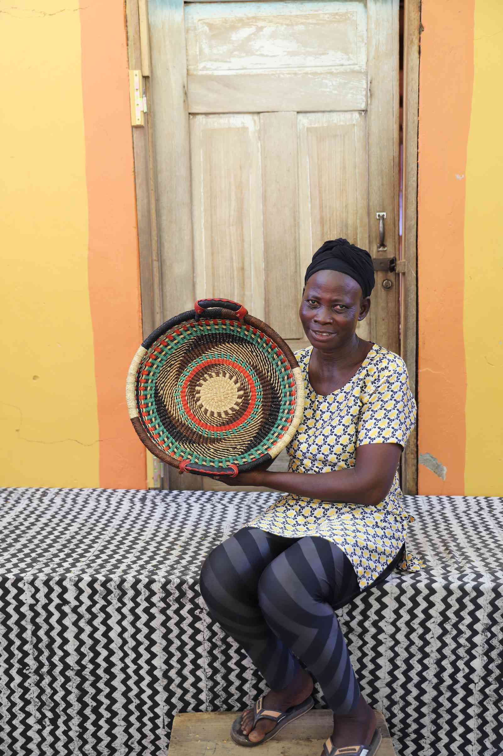 Handwoven Bolga wall tray disc with concentric pink, red and teal bands and yellow centre, held by a seated woman.