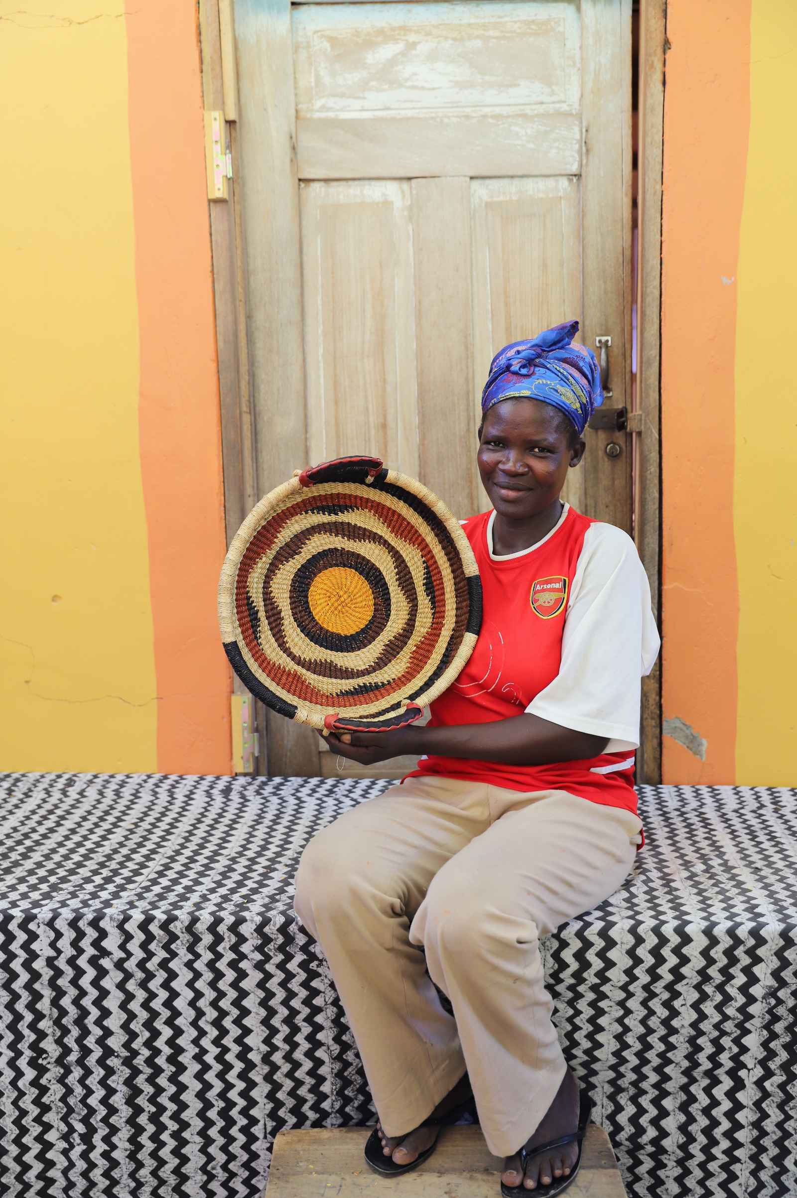 Handwoven Bolga wall tray disc with red, black and orange concentric bands, held by a seated woman.