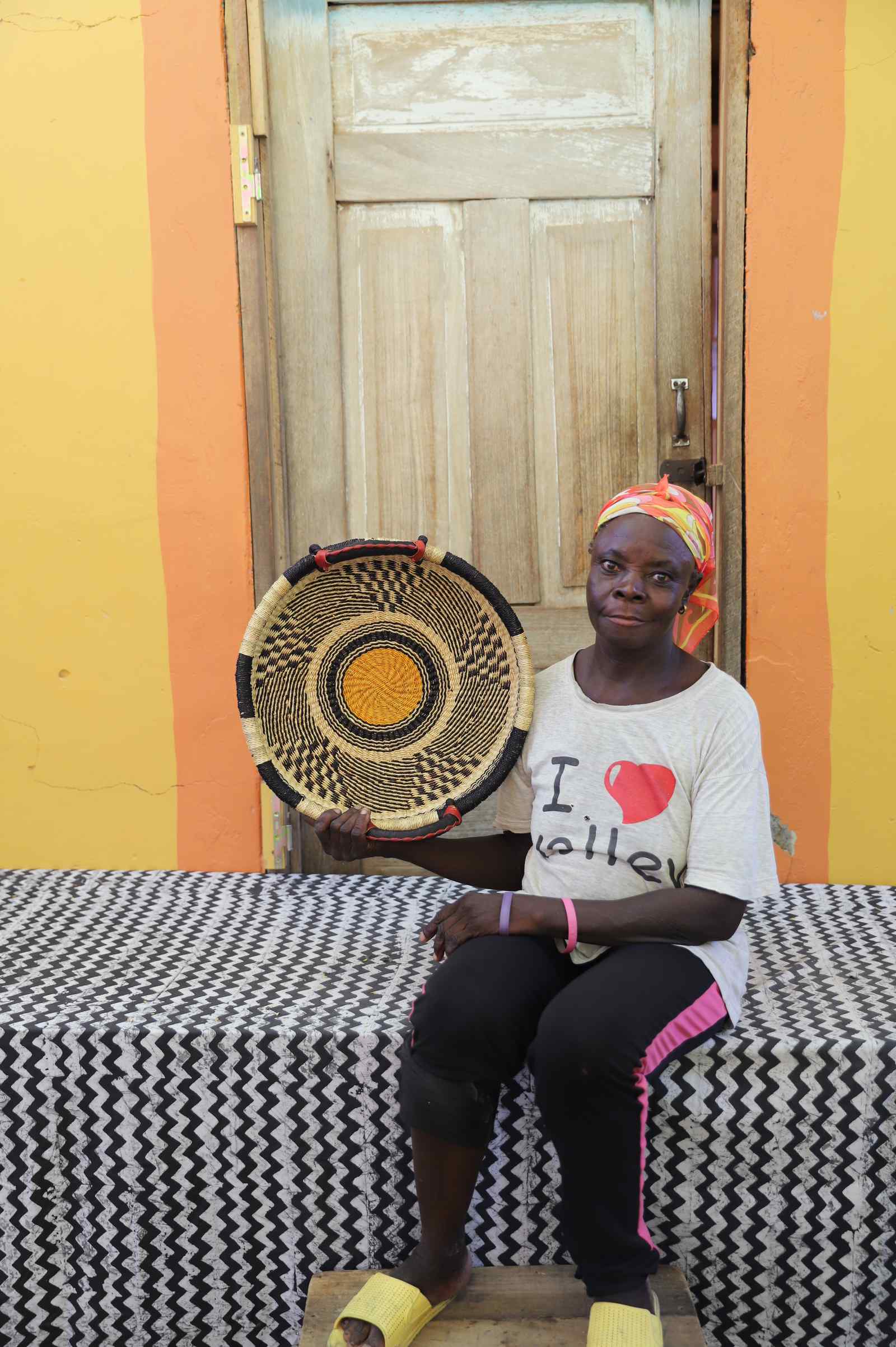 Handwoven Bolga wall tray disc in natural straw with a yellow-orange bullseye centre, held by a seated woman.