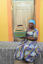 Handwoven round Bolga shopping basket in green and teal, held by a seated woman in a patterned dress.