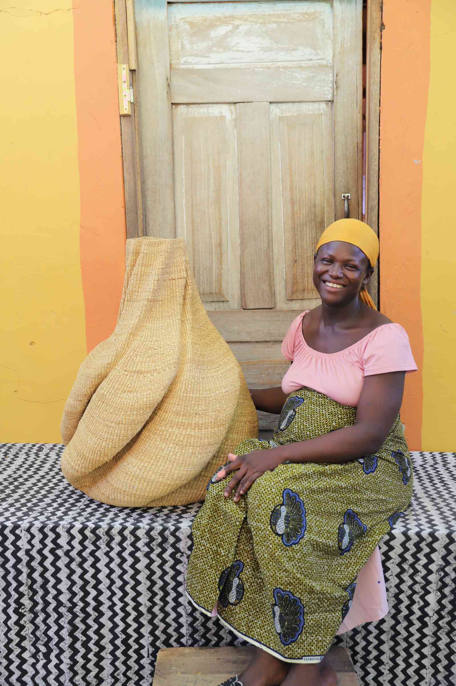 Medium handwoven Bolga art basket in natural straw with a sculpted lobed form, beside a seated woman in pink.