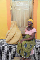 Medium handwoven Bolga art basket in natural straw with a sculpted lobed form, beside a seated woman in pink.