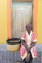 Small handwoven Bolga storage tub in plain natural straw, beside a seated woman in red and white.