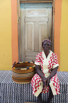 Small handwoven Bolga storage tub in natural with fine red stripes, beside a seated woman in red and white.