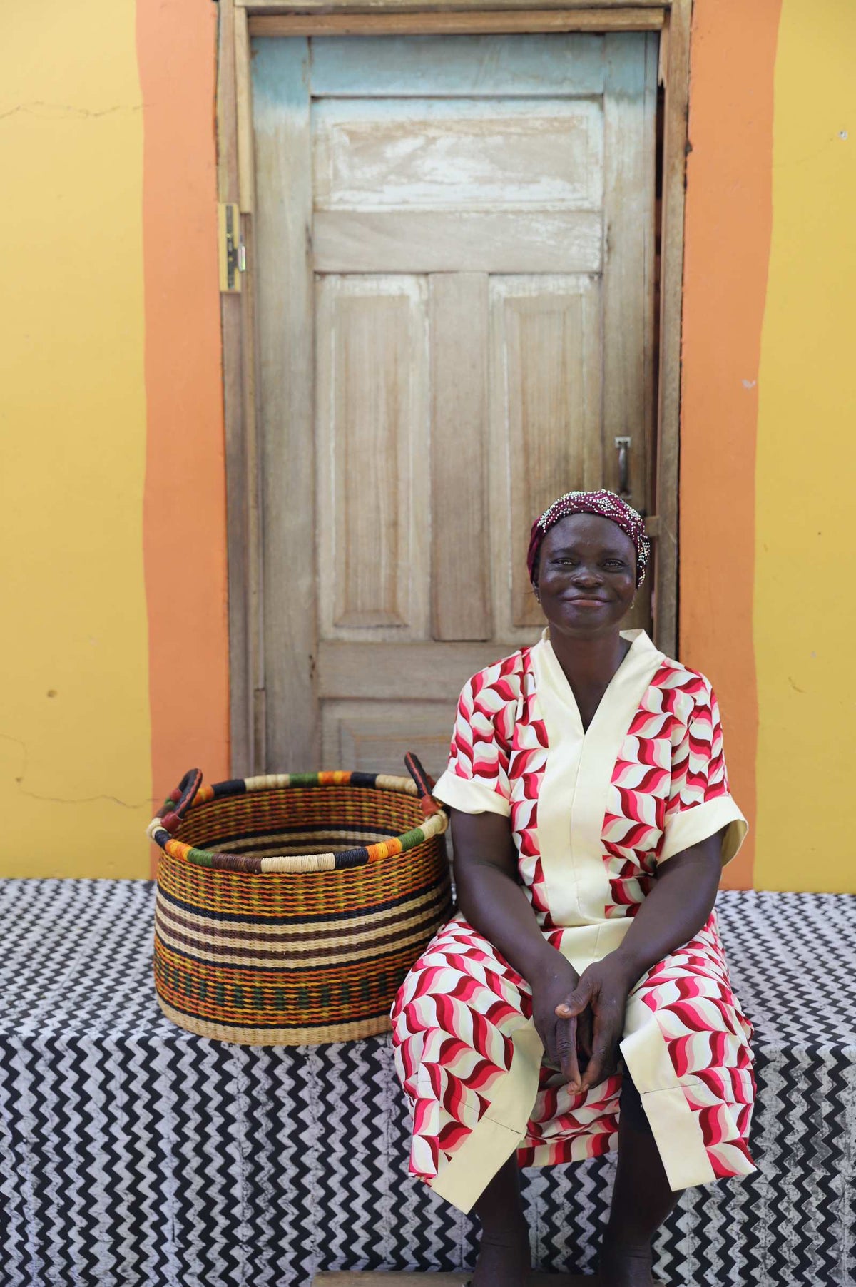 Small handwoven Bolga storage tub in natural with fine red stripes, beside a seated woman in red and white.