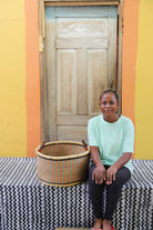 Small handwoven Bolga storage tub in natural with orange and green bands, beside a seated woman in green.