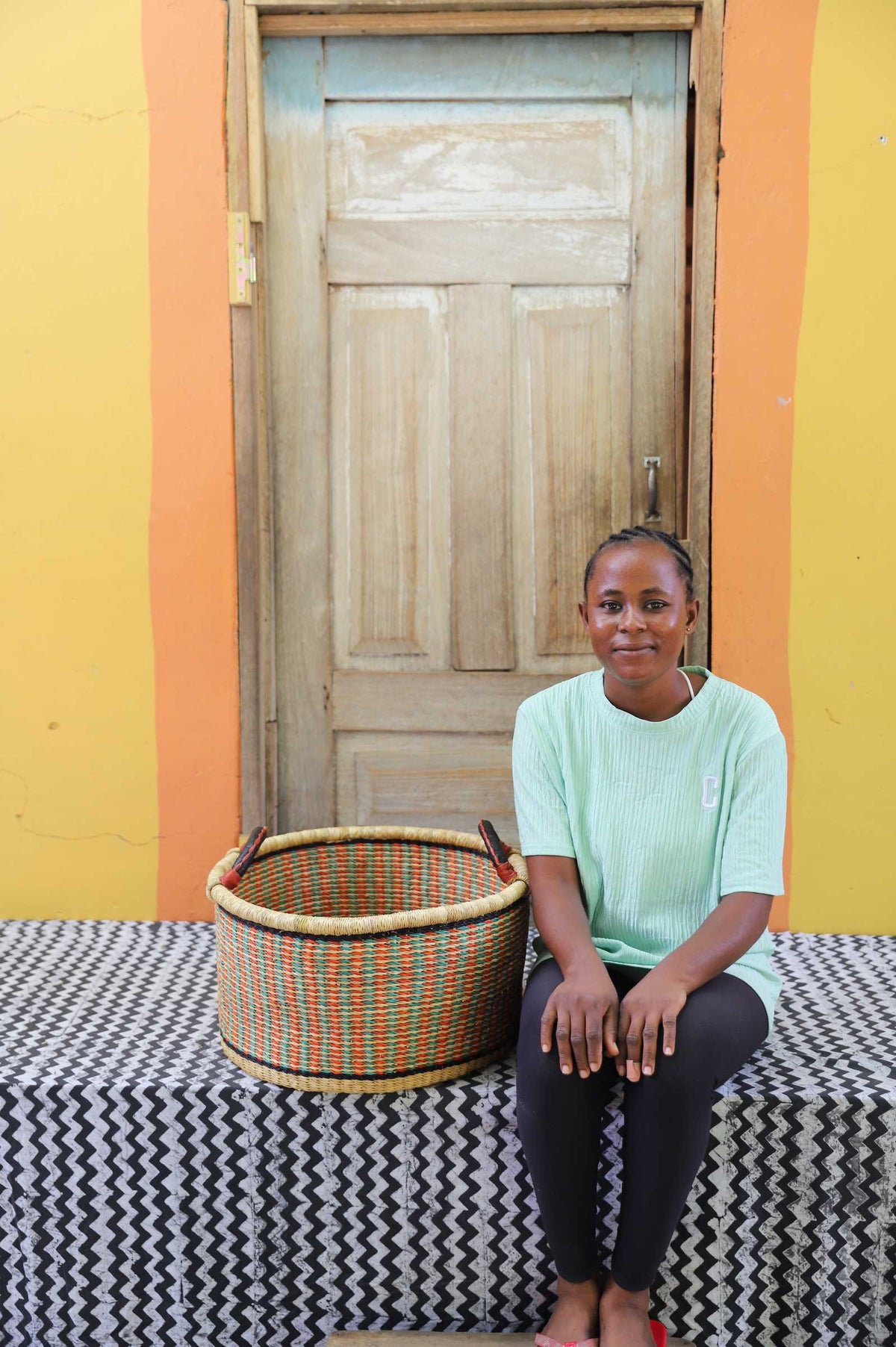 Small handwoven Bolga storage tub in natural with orange and green bands, beside a seated woman in green.