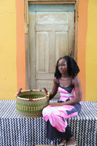 Small handwoven Bolga storage tub in dark green, beside a seated woman in pink and black.