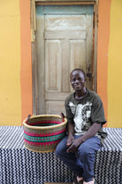 Small handwoven Bolga storage tub in natural with red and green bands, beside a seated man in patterned shirt.