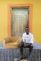 Large handwoven Bolga pet bed in plain natural straw, beside a seated man in white.