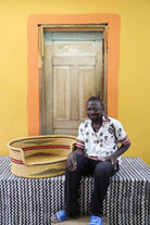 Large handwoven Bolga pet bed in natural with fine red stripes, beside a seated man in a printed shirt.