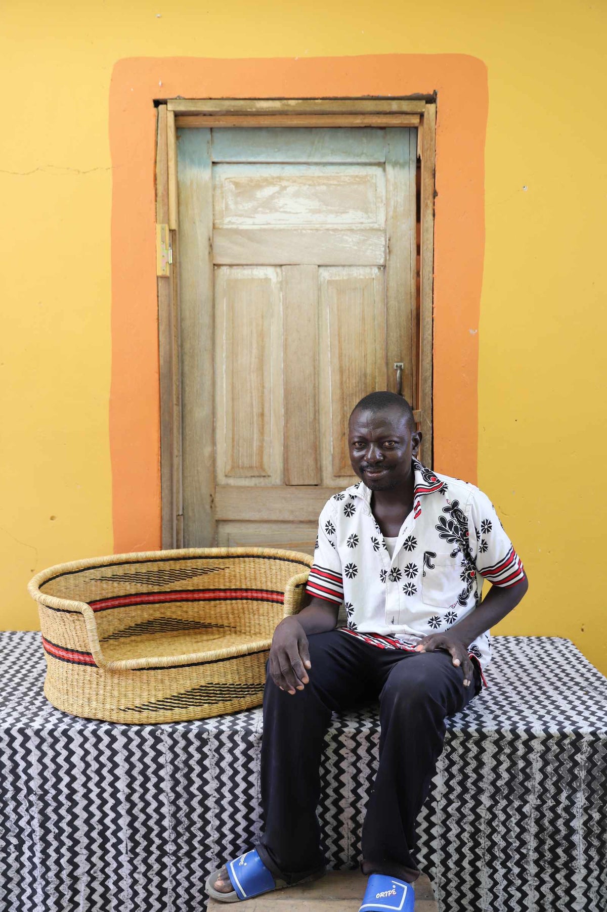 Large handwoven Bolga pet bed in natural with fine red stripes, beside a seated man in a printed shirt.