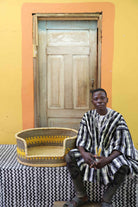 Medium handwoven Bolga pet bed in natural with yellow stripes, beside a seated man in stripes.