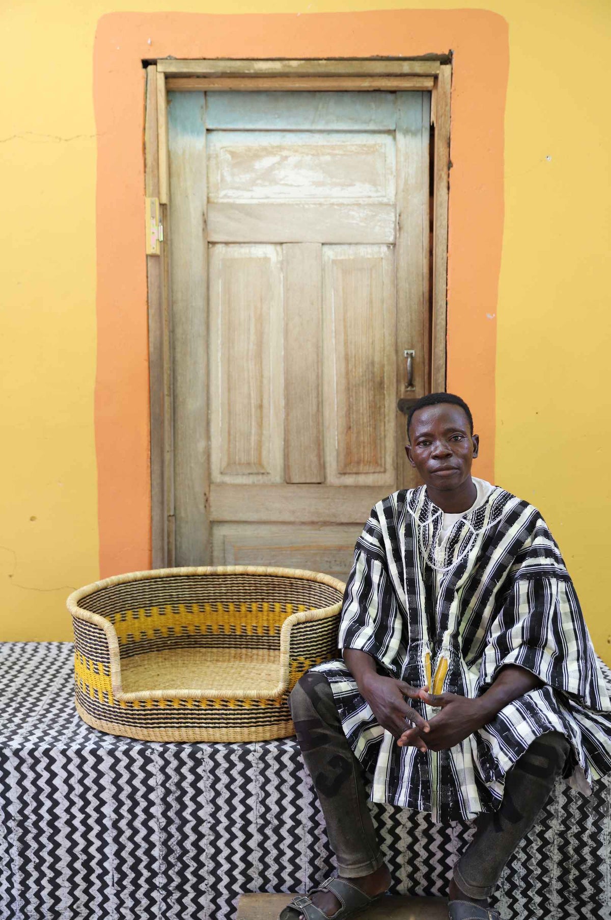 Medium handwoven Bolga pet bed in natural with yellow stripes, beside a seated man in stripes.