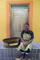 Small handwoven Bolga pet bed in dark brown straw with a natural woven interior, beside a seated woman in green.